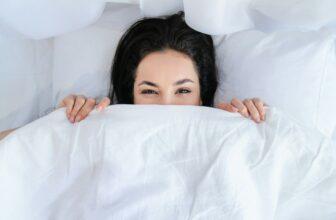 Smiling young pretty woman hiding under the blanket in her bed in the morning